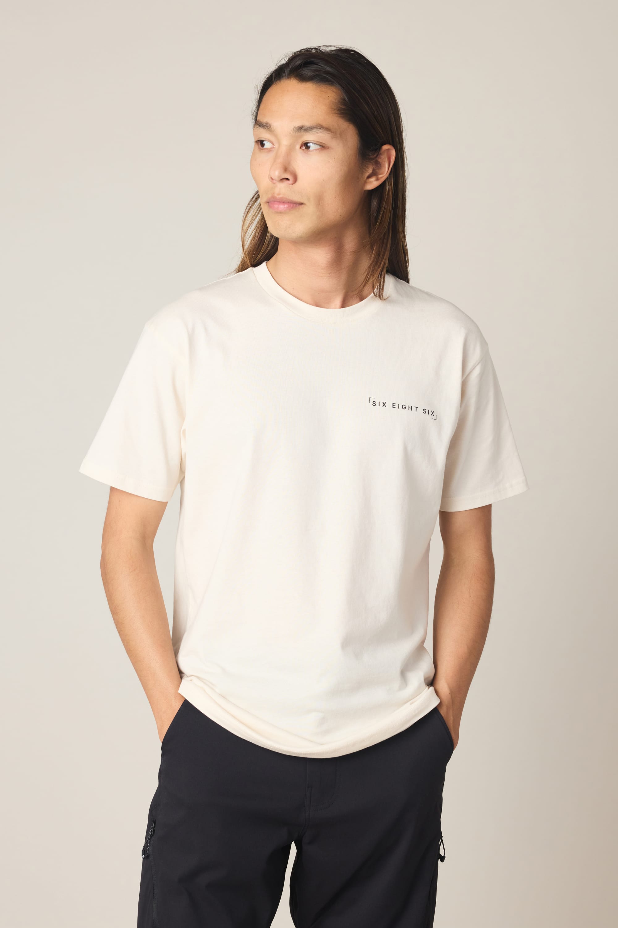 A person with long hair stands against a plain background, hands in pockets, wearing the 686 24/7 Authentic SS Tee by 686—a cream men's cotton t-shirt with small chest text—paired with dark pants, looking slightly to the side.