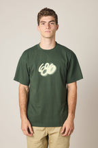 A young man with short curly hair wears the 686 Spray Relaxed SS Tee by 686—a dark green 100% cotton men’s t-shirt with a white graffiti-style “666” chest design—and light khaki pants, standing before a plain light background.