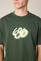 A person wearing the 686 Spray Relaxed SS Tee—a dark green men’s t-shirt featuring a white, spray-painted 686 logo. Only the lower face and upper torso are shown, emphasizing its 100% cotton fabric.