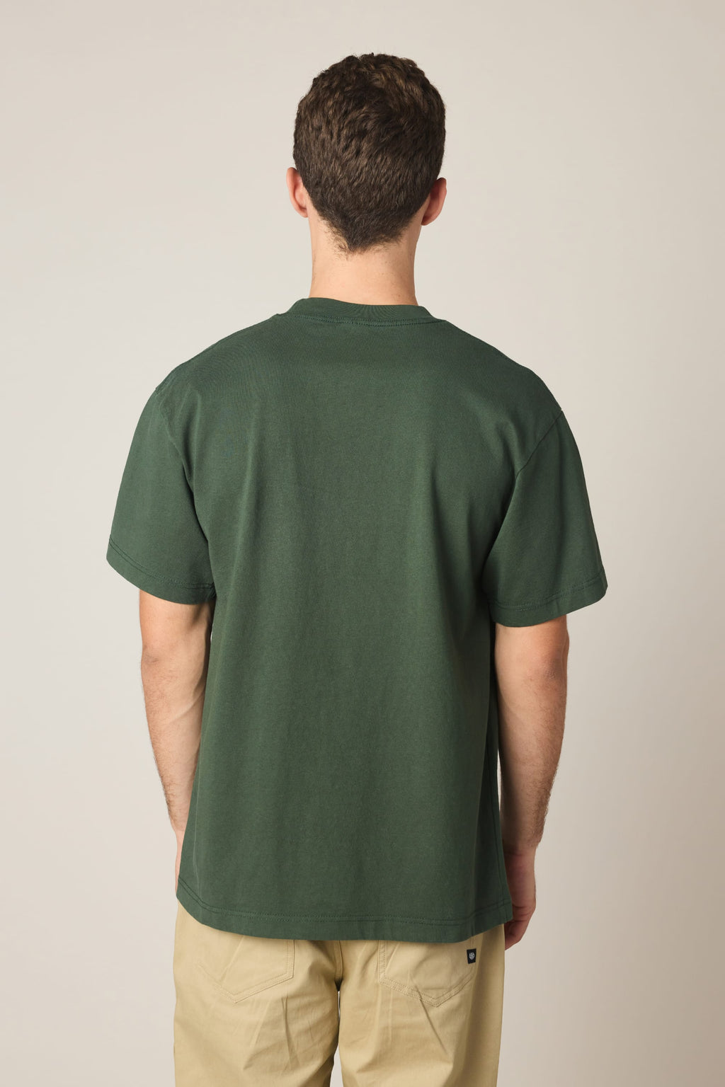 A person with short brown hair stands with their back to the camera, wearing the 686 Spray Relaxed SS Tee by 686 in dark green and light beige pants, against a neutral background.