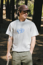 A man in green pants, a brown cap, and the 686 Spray Relaxed SS Tee by 686—a light gray, 100% cotton men's t-shirt with a blue design—stands in a wooded area holding a stick and looking to the side.