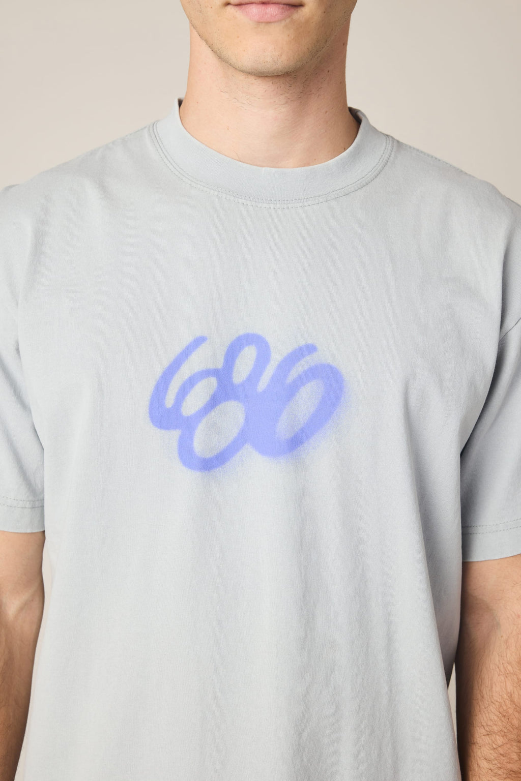 A person wears the 686 Spray Relaxed SS Tee by 686, a light gray men's t-shirt featuring a blue graffiti-style "686" on the chest. Made from soft 100% cotton, it offers comfort; the wearer’s face is partially out of frame.