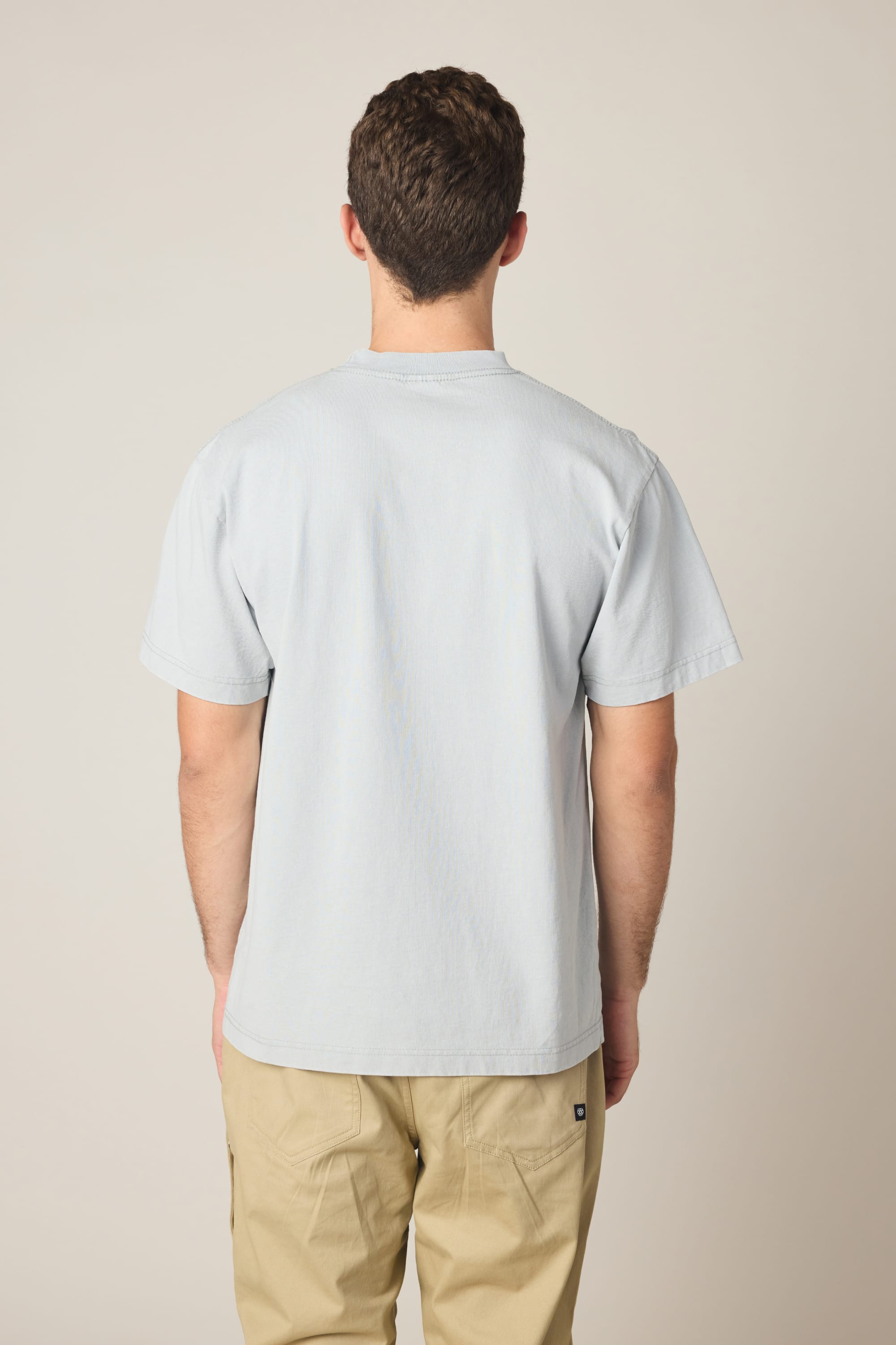 A person with short brown hair is seen from behind, wearing the 686 Spray Relaxed SS Tee by 686 in light gray and beige pants, set against a plain light background.