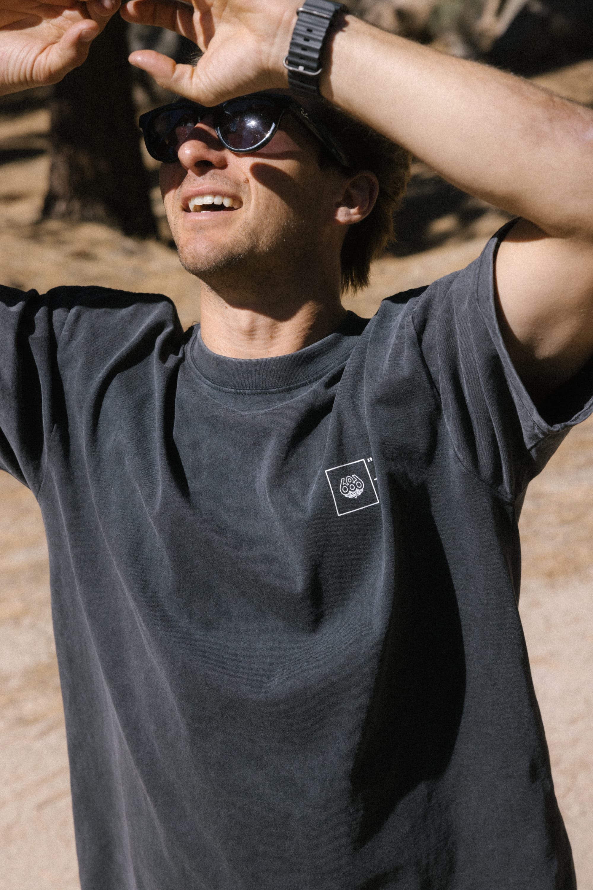 Wearing the 686 Engineered Relaxed SS Tee, a person in sunglasses shields their eyes from the sun with one arm while standing outdoors on a bright, sunny day.