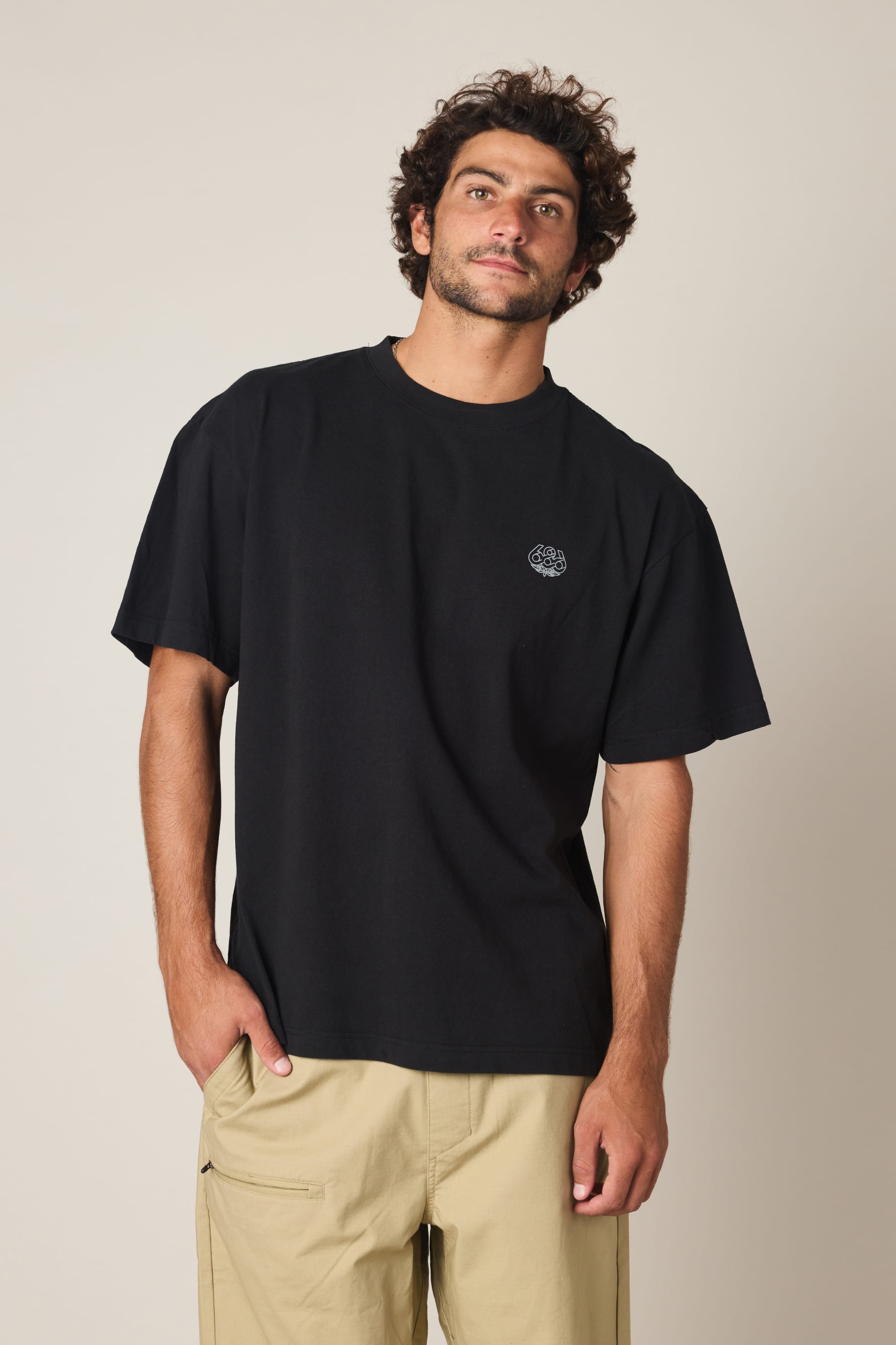 A man with curly hair and a beard wears the 686 Engineered Relaxed SS Tee and light beige shorts, standing against a plain light background with one hand in his pocket and a slight smile.