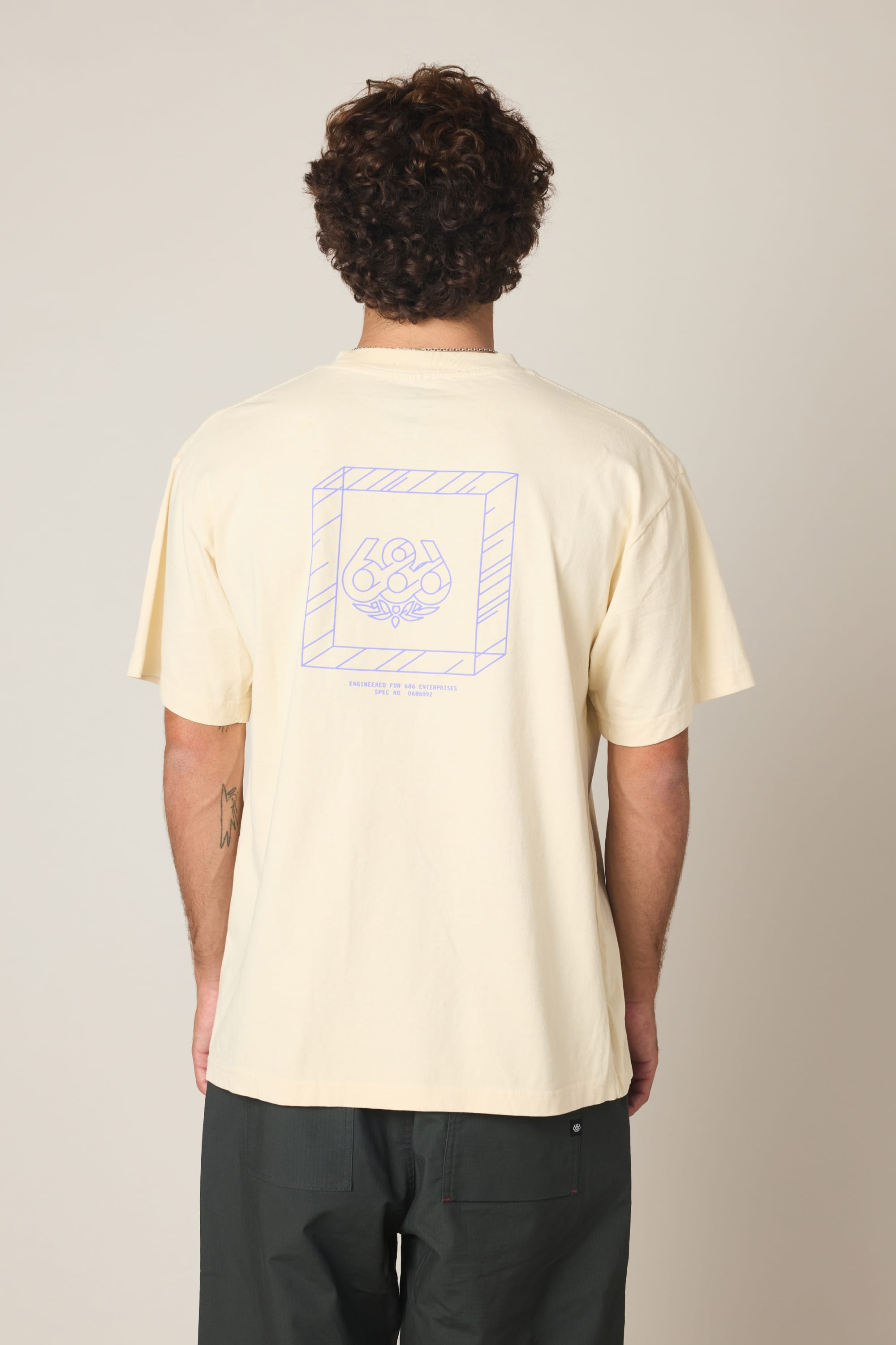 A person with curly hair is seen from behind wearing the 686 Engineered Relaxed SS Tee—a cream men's 100% cotton shirt with a purple geometric graphic and “686” on the back—paired with dark pants against a plain, light background.