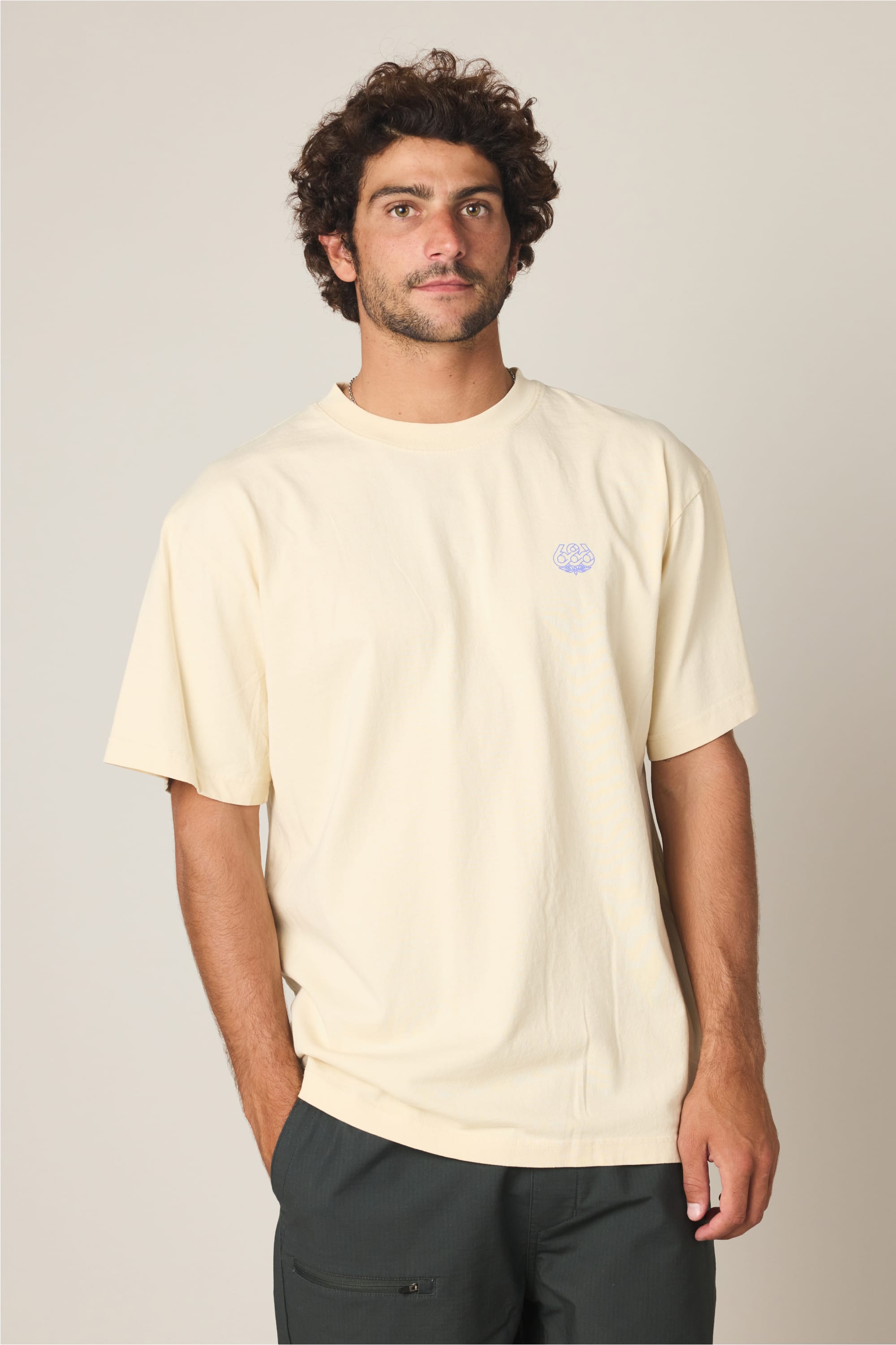 A man with curly brown hair and a beard stands against a plain background, wearing the 686 Engineered Relaxed SS Tee by 686—a loose, light yellow 100% cotton men's t-shirt with a small logo on the chest—and dark pants. He looks slightly off to the side.