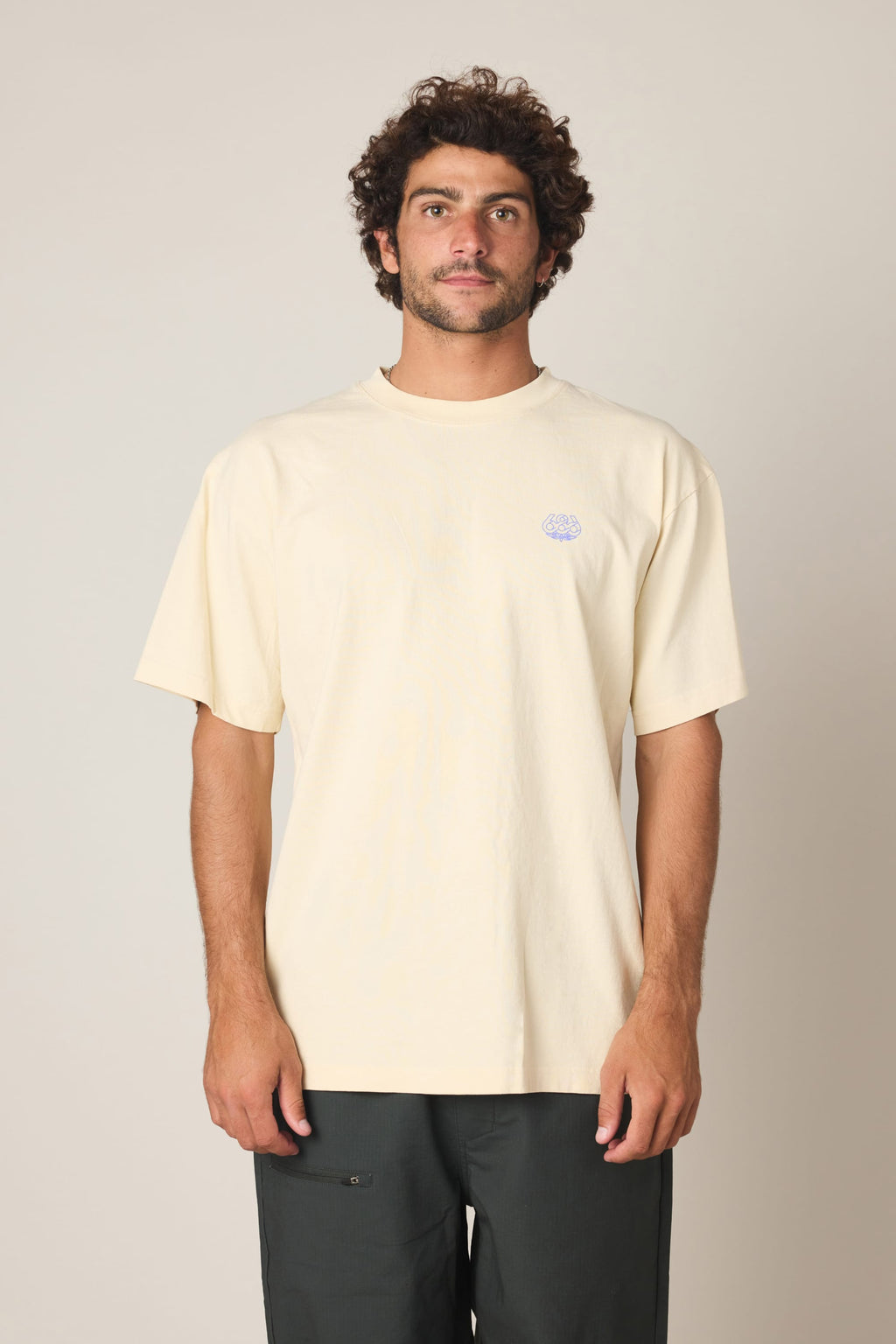 A man with curly dark hair and a beard stands facing forward, wearing the 686 Engineered Relaxed SS Tee in pale yellow with a small circular logo on the left chest, paired with dark pants, against a plain light background.