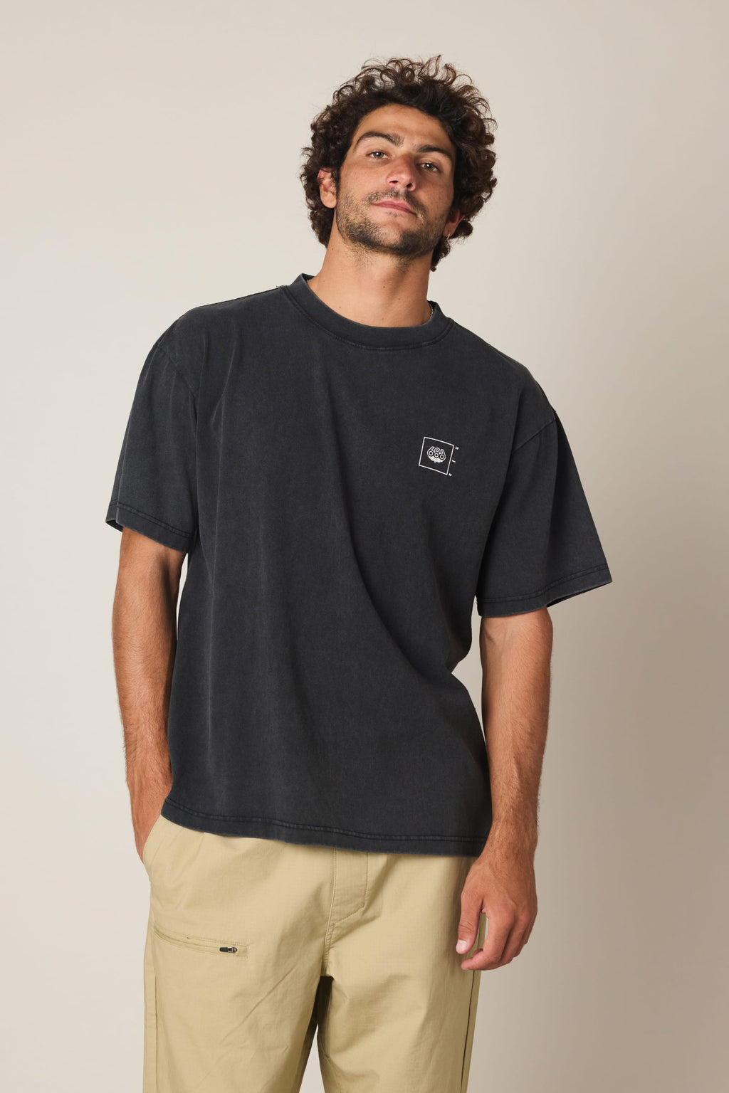 A man with curly hair and a beard is wearing the 686 Peaks Relaxed SS Tee by 686, a loose-fitting 100% cotton men’s t-shirt with a small white chest logo, paired with beige pants against a plain light background.