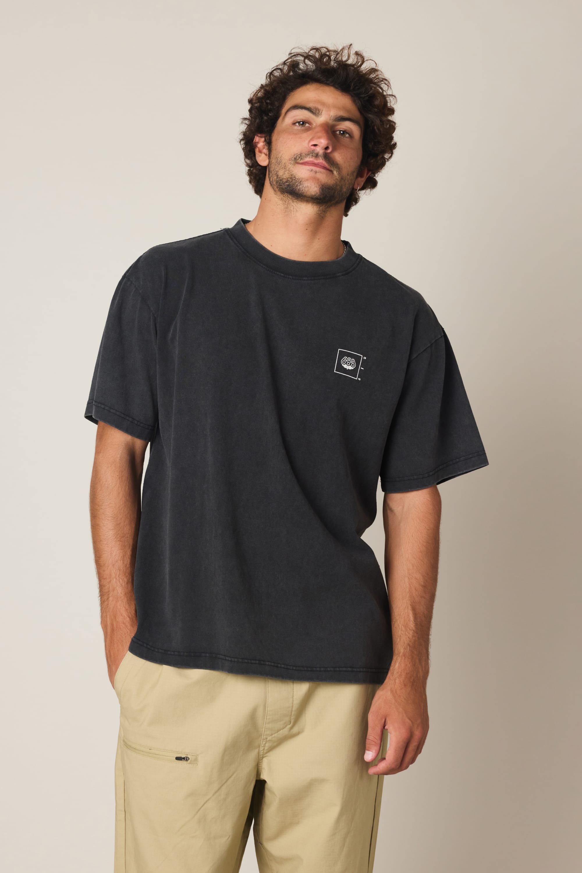 A man with curly hair and a beard is wearing the 686 Peaks Relaxed SS Tee by 686, a loose-fitting 100% cotton men’s t-shirt with a small white chest logo, paired with beige pants against a plain light background.