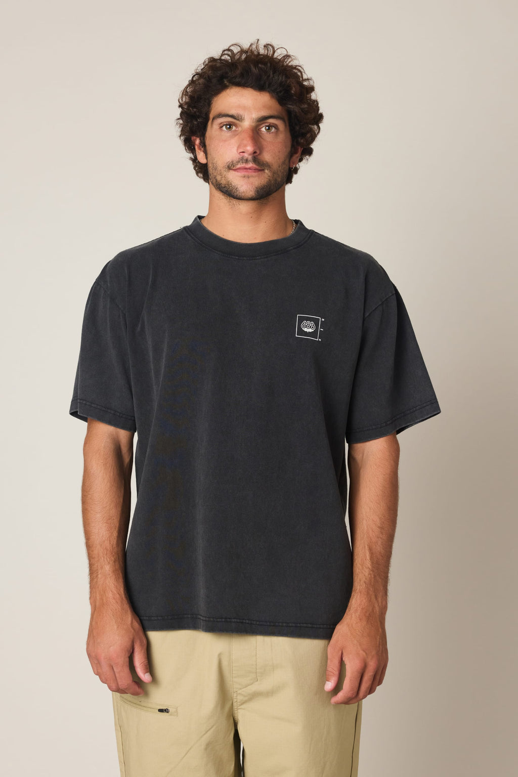 A man with curly brown hair and a beard wears the 686 Peaks Relaxed SS Tee—a loose-fitting, faded black 100% cotton men's t-shirt with a small white logo on the chest—paired with beige shorts against a plain light background.