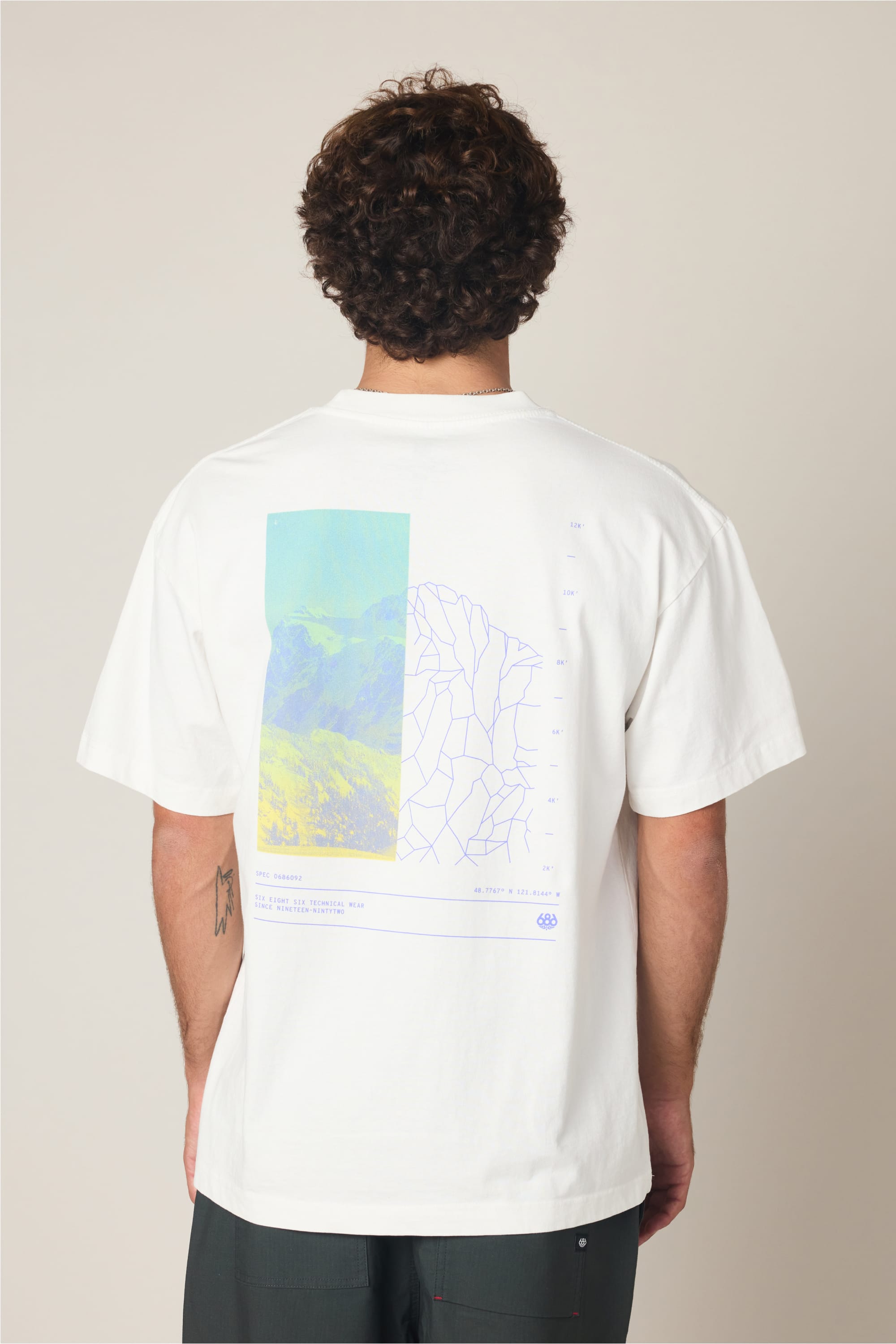A person with curly hair faces away, wearing the 686 Peaks Relaxed SS Tee by 686—a 100% cotton men’s T-shirt with a colorful mountain and abstract line art graphic on the back in blue, green, and purple tones.