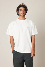 A man with curly hair and a beard stands against a light background, wearing the 686 Peaks Relaxed SS Tee by 686 and dark pants. His right hand is in his pocket as he gazes slightly past the camera with a neutral expression.