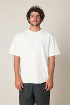 A man with curly hair and a beard stands facing forward, wearing the 686 Peaks Relaxed SS Tee by 686—a plain white oversized men's t-shirt made from 100% cotton with a small chest logo—paired with black pants against a neutral background.