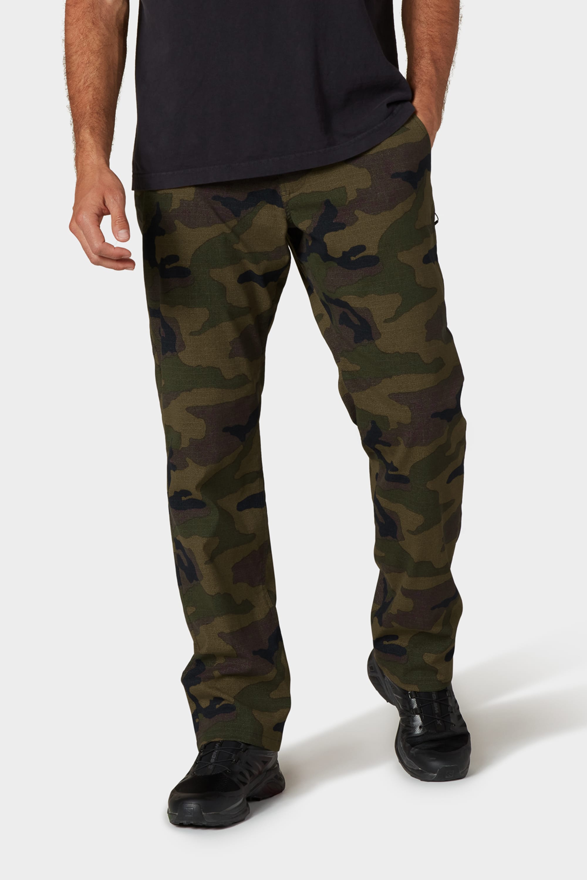 A man, visible from the shoulders down, stands against a white background wearing a black t-shirt and 686 Men's Everywhere® Merino-Lined Pant - Relaxed Fit in green camo, paired with black sneakers. His hands rest relaxed at his sides.