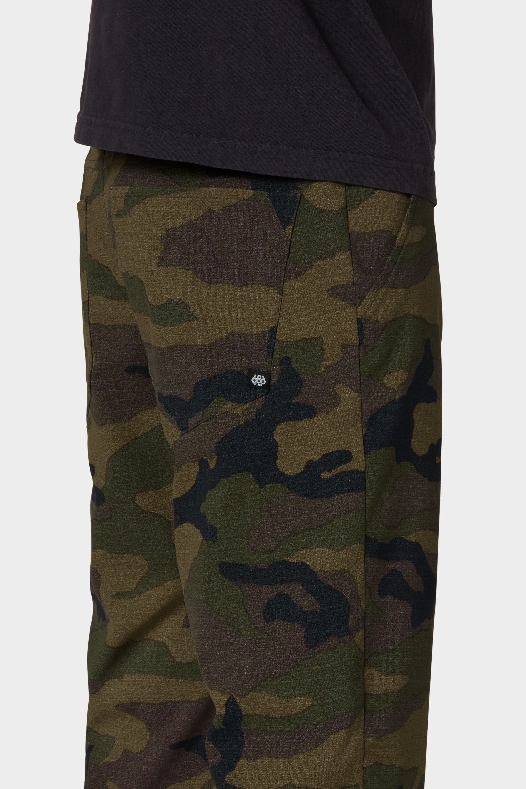 A close-up side view of a person wearing the 686 Men's Everywhere® Merino-Lined Pant - Relaxed Fit in green and brown camouflage, paired with a black shirt and a stylish insulated jacket featuring a small round logo on the pocket.