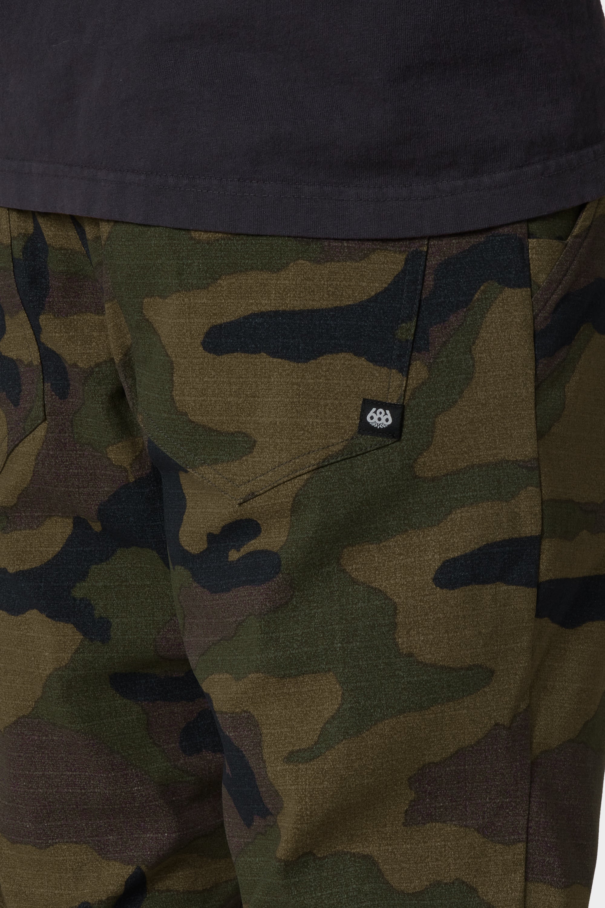 A person shown from the waist down wears 686 Men's Everywhere® Merino-Lined Pant - Slim Fit in green and brown camo and a black shirt. The pants, branded by 686, keep you warm in cold weather and feature a small black and white logo tag near the pocket.