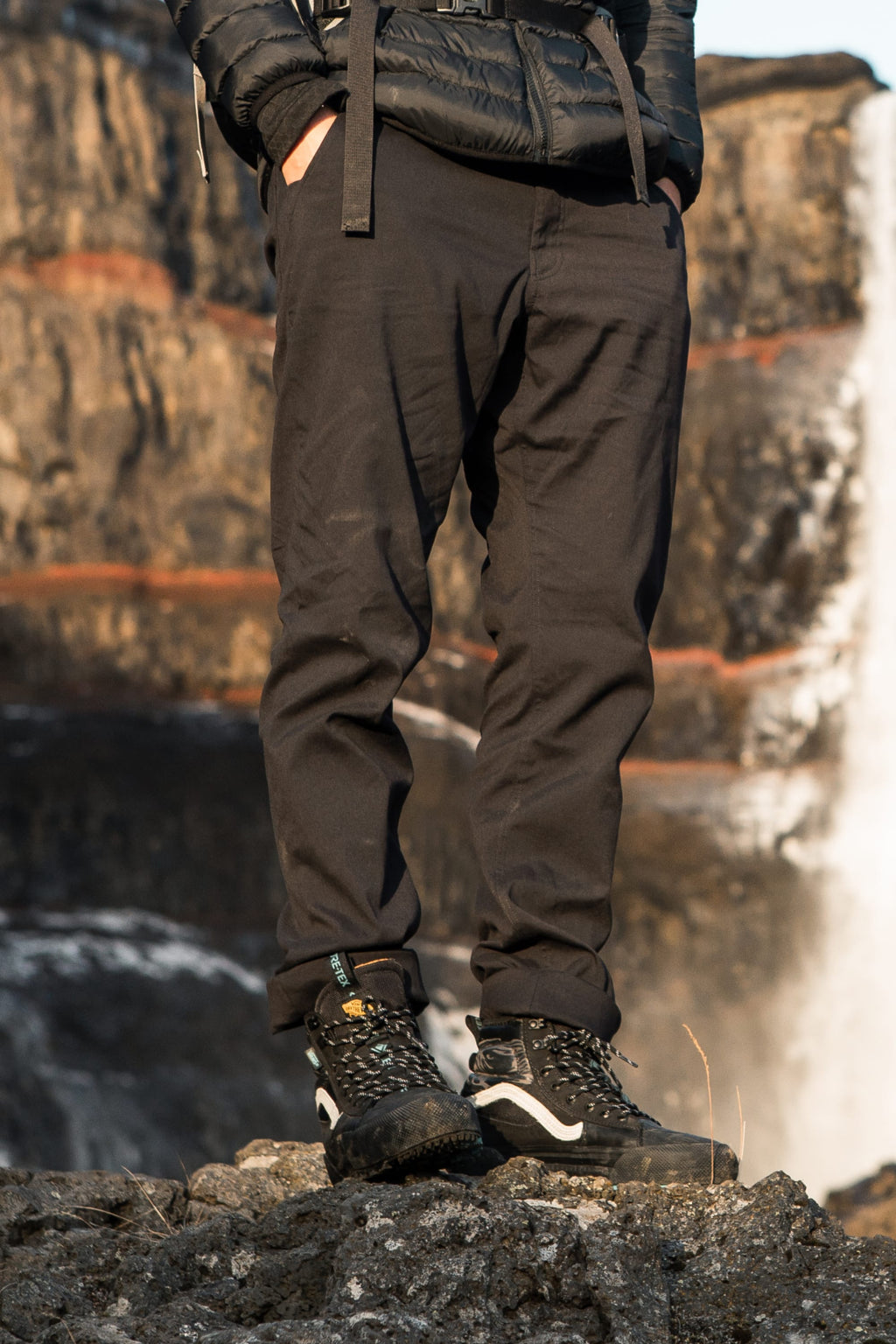 Wearing 686 Men's Everywhere® Merino-Lined Pant - Relaxed Fit, a sleek insulated jacket, and black hiking boots, a person stands on rocky terrain outdoors with a blurred waterfall and cliffs in the background.
