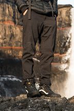 Wearing 686 Men's Everywhere® Merino-Lined Pant - Relaxed Fit, a sleek insulated jacket, and black hiking boots, a person stands on rocky terrain outdoors with a blurred waterfall and cliffs in the background.
