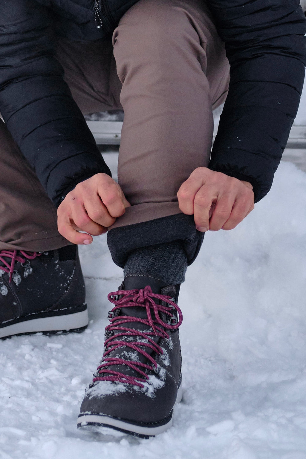 Wearing the 686 Men's Everywhere® Merino-Lined Pant - Relaxed Fit, a person rolls up a pant leg to reveal thermal leggings, sitting in the snow with dark winter boots, pink laces, and a black jacket for added warmth.