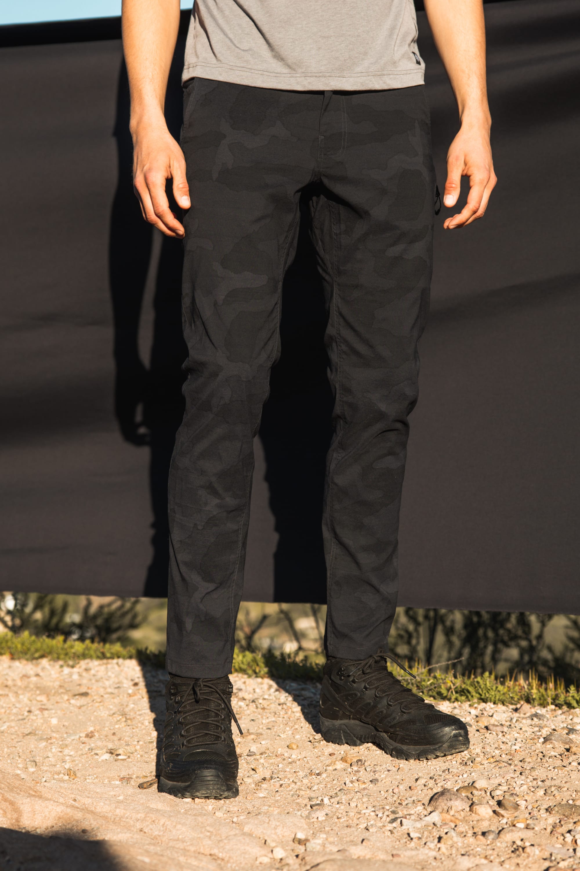 A person stands outdoors on gravel, wearing the 686 Mens Everywhere Featherlight Chino Pant - Slim Fit in black camo, paired with black boots and a gray shirt. A black cloth backdrop and blurred scenery highlight these lightweight pants by 686 for warm-weather adventures.