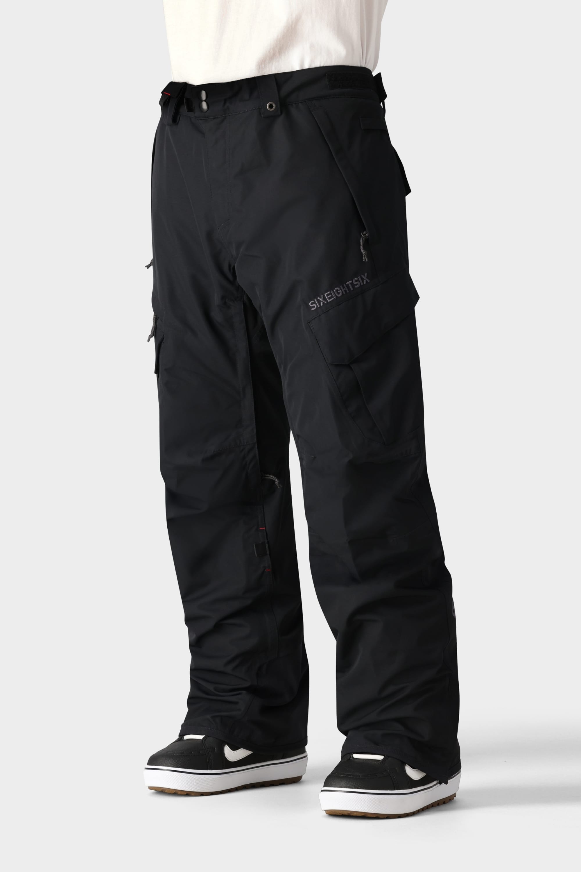 A person wears 686 Men's SMARTY® 3-in-1 Cargo Pant—versatile black cargo pants with multiple pockets and a removable sweatpant liner—paired with black sneakers with white soles, standing against a light gray background. Upper body not shown.