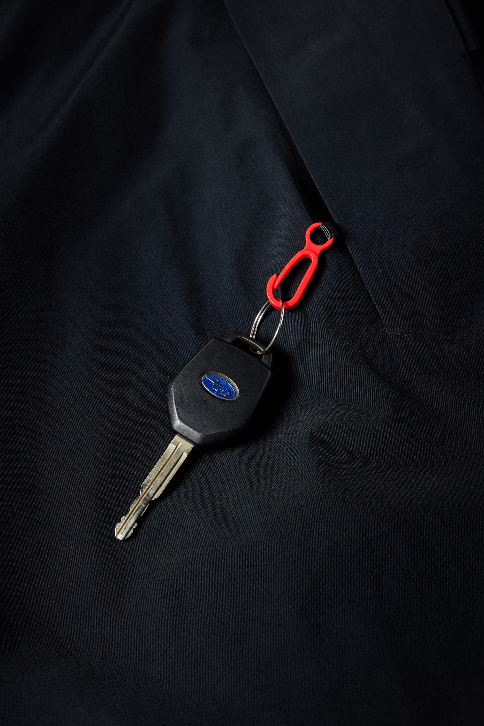 A metal car key with a black plastic head and blue oval logo is attached to a red clip, resting on dark fabric—likely the 686 Men's SMARTY® 3-in-1 Cargo Pant by 686, which features a removable sweatpant liner for versatile wear.