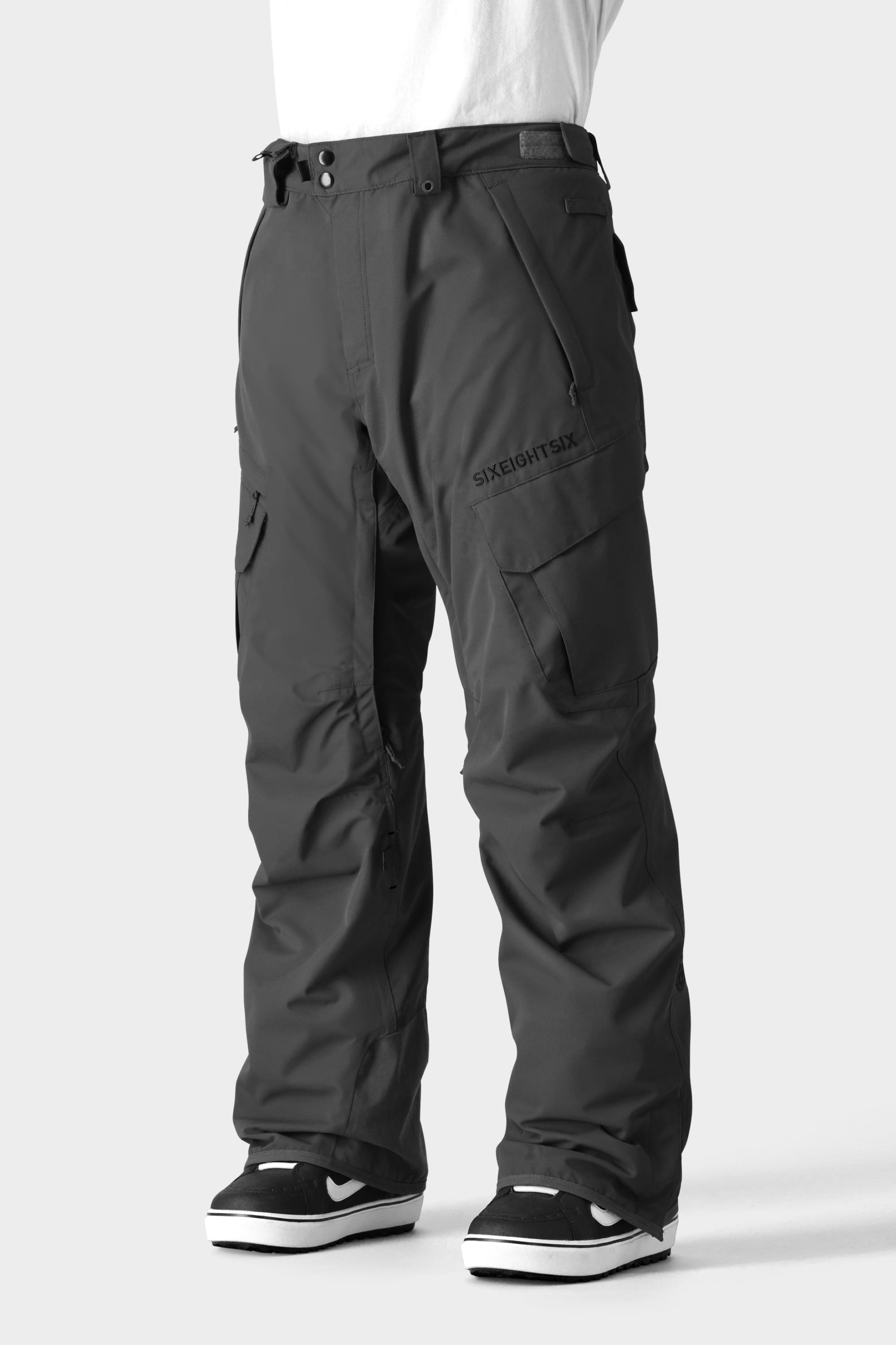 A person wears the 686 Men's SMARTY® 3-in-1 Cargo Pant by 686, featuring a removable liner and zipper details, paired with black-and-white sneakers against a light gray background.