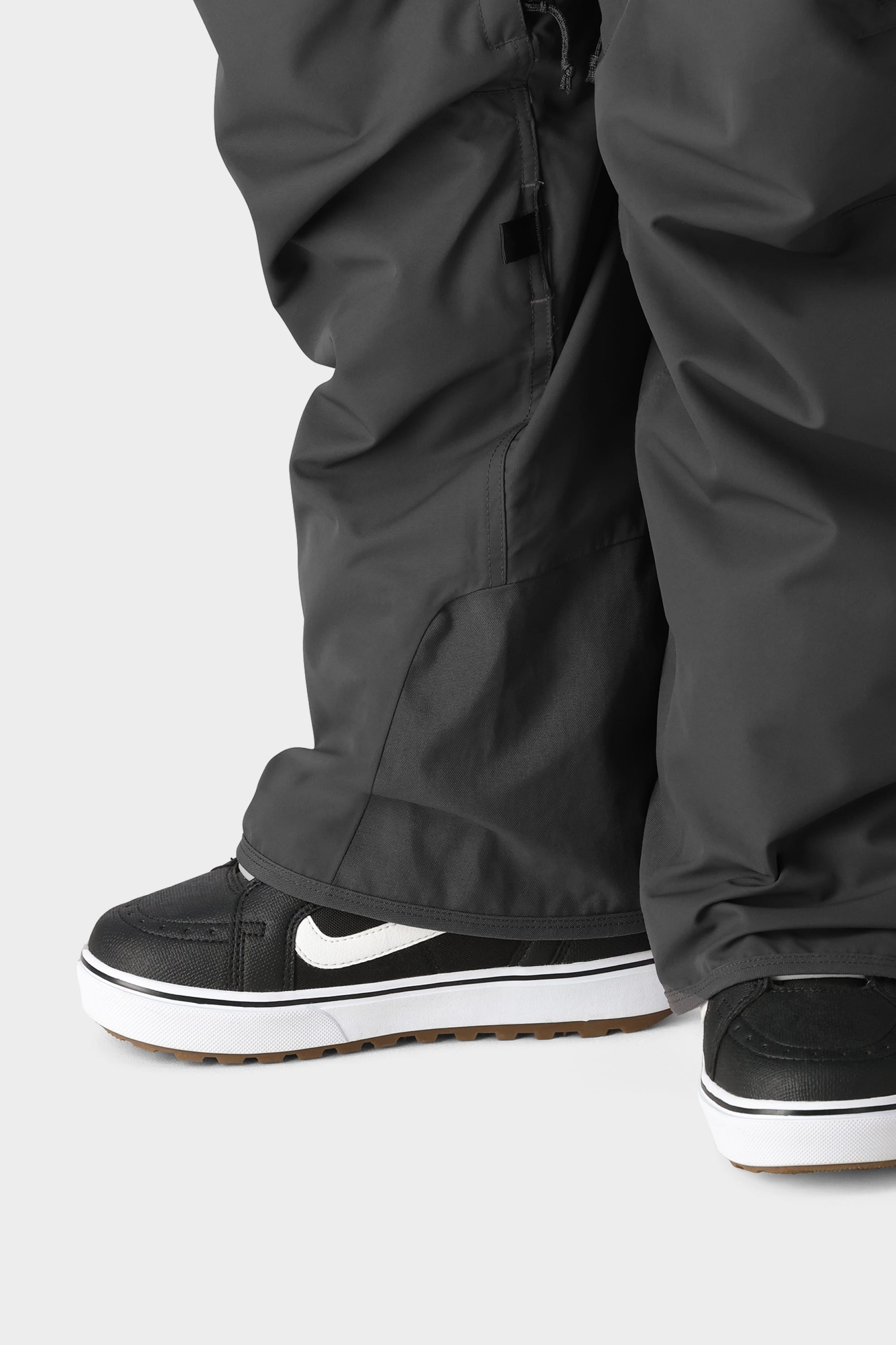 A close-up of a person wearing the 686 Men's SMARTY® 3-in-1 Cargo Pant with removable sweatpant liner, paired with black and white Nike sneakers with gum soles, standing on a light gray background—offering three ways to wear.