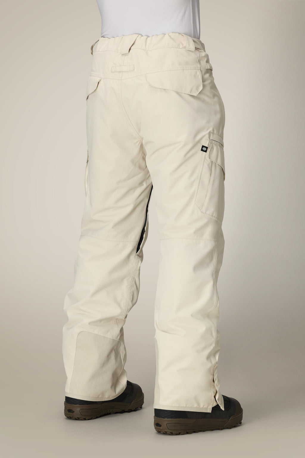 A person in 686 Men's SMARTY® 3-in-1 Cargo Pant with black boots faces away, showing the back and side pockets. These off-white pants by 686 have a removable sweatpant liner, offering three ways to wear them against a plain light background.