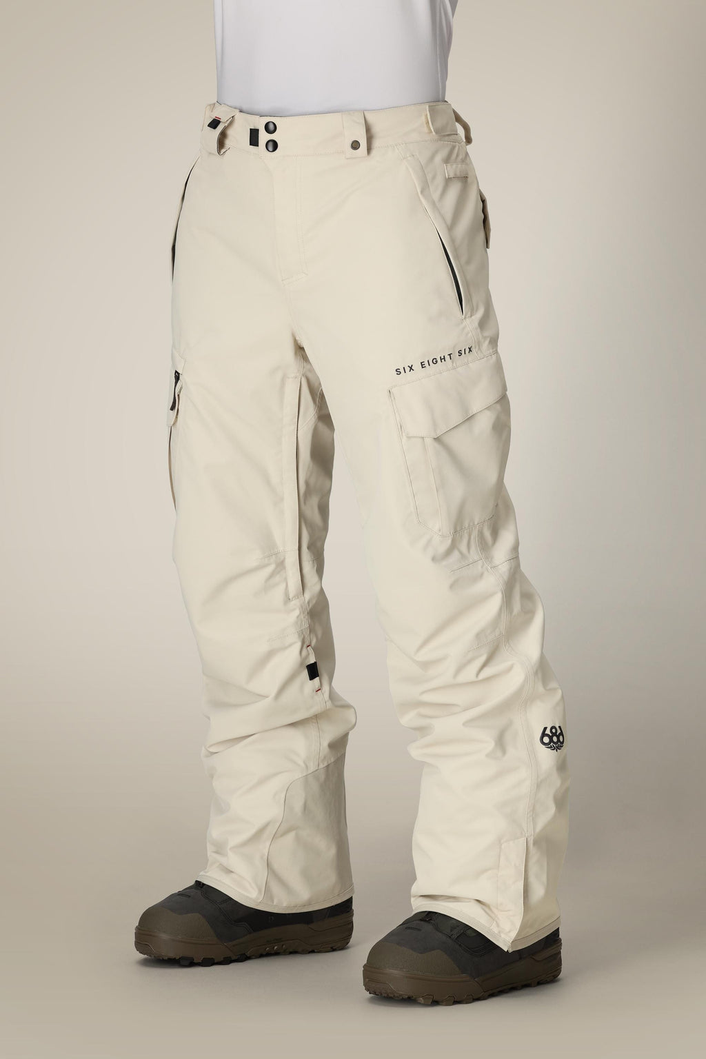 A person wears 686 Men's SMARTY® 3-in-1 Cargo Pant by 686 in off-white, featuring multiple pockets, black zippers, branded text on the thigh, and black boots, against a plain background. The pants offer three versatile wearing options.