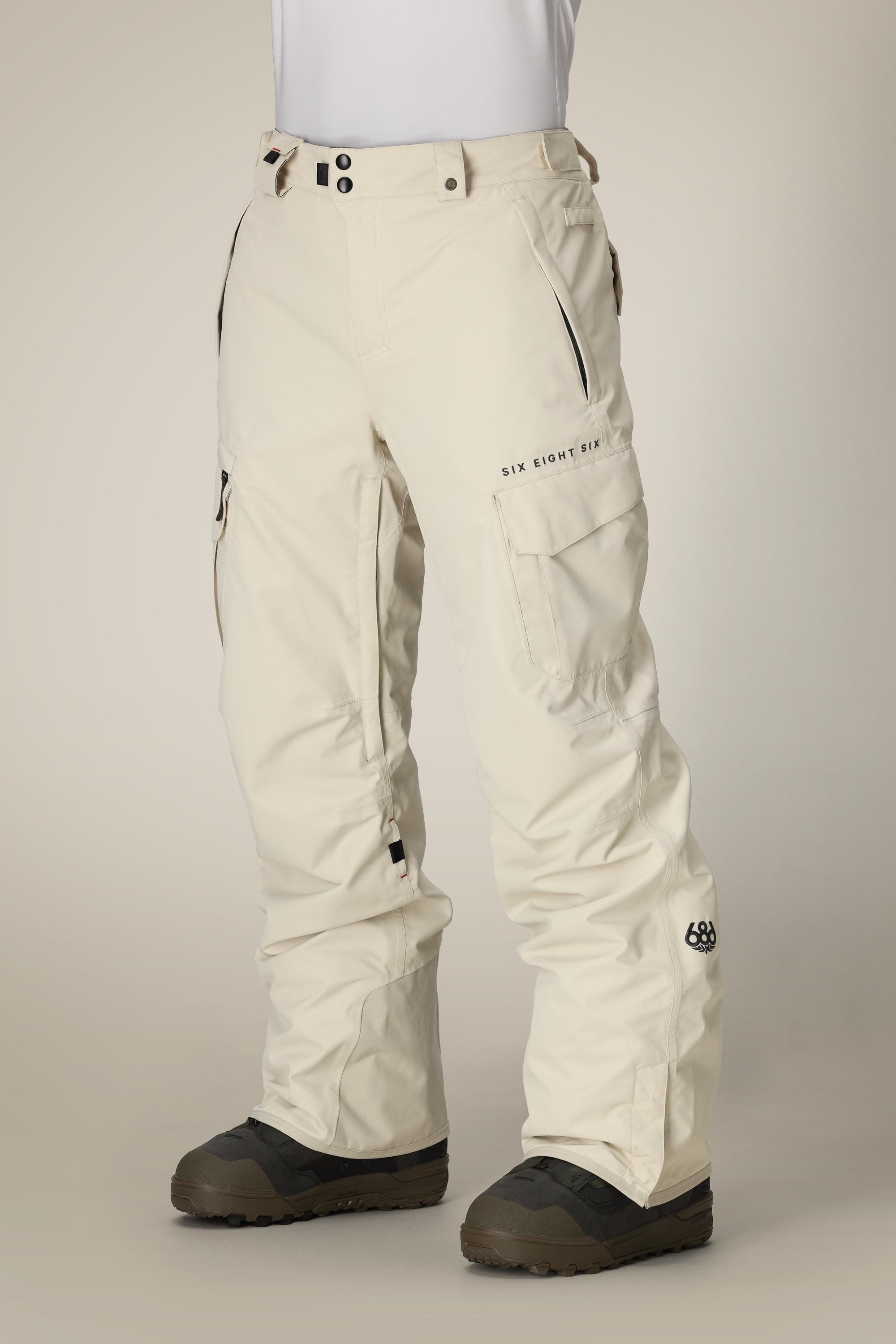 A person wears 686 Men's SMARTY® 3-in-1 Cargo Pant by 686 in off-white, featuring multiple pockets, black zippers, branded text on the thigh, and black boots, against a plain background. The pants offer three versatile wearing options.