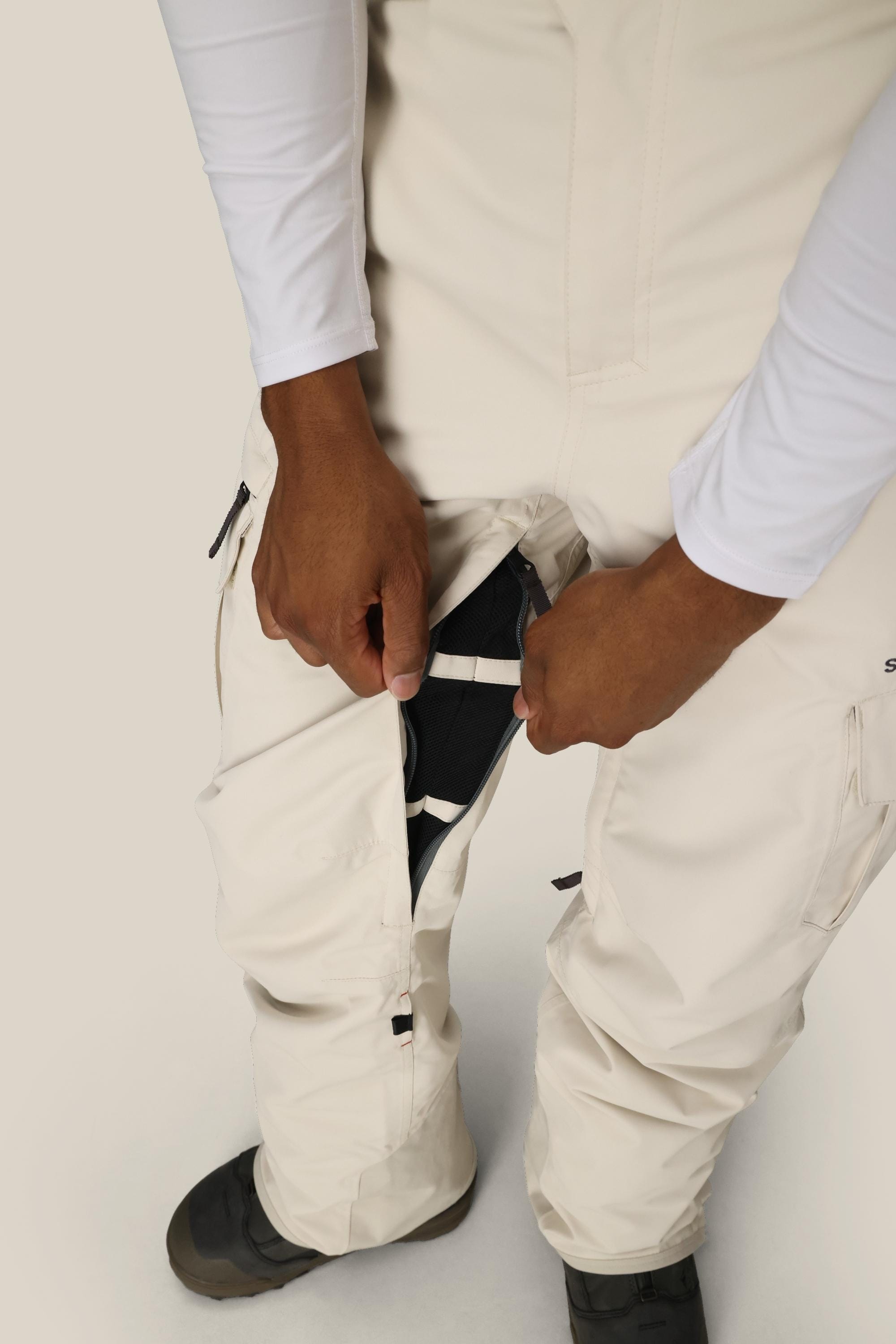A person adjusts the zipper on the 686 Men's SMARTY® 3-in-1 Cargo Pant, revealing its black removable liner. The off-white pants, paired with a white long-sleeve shirt, showcase three versatile wear options from brand 686.