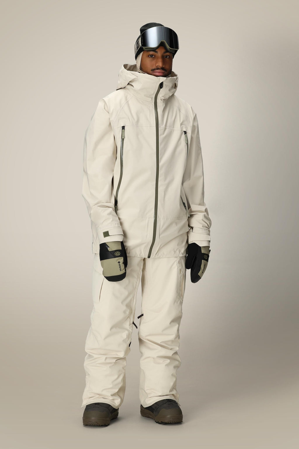 A person wears a cream ski jacket and 686 Men's SMARTY® 3-in-1 Cargo Pant by 686, featuring a removable sweatpant liner, plus black gloves, brown boots, and ski goggles, standing against a plain light background.