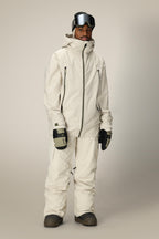A person wears a cream ski jacket and 686 Men's SMARTY® 3-in-1 Cargo Pant by 686, featuring a removable sweatpant liner, plus black gloves, brown boots, and ski goggles, standing against a plain light background.