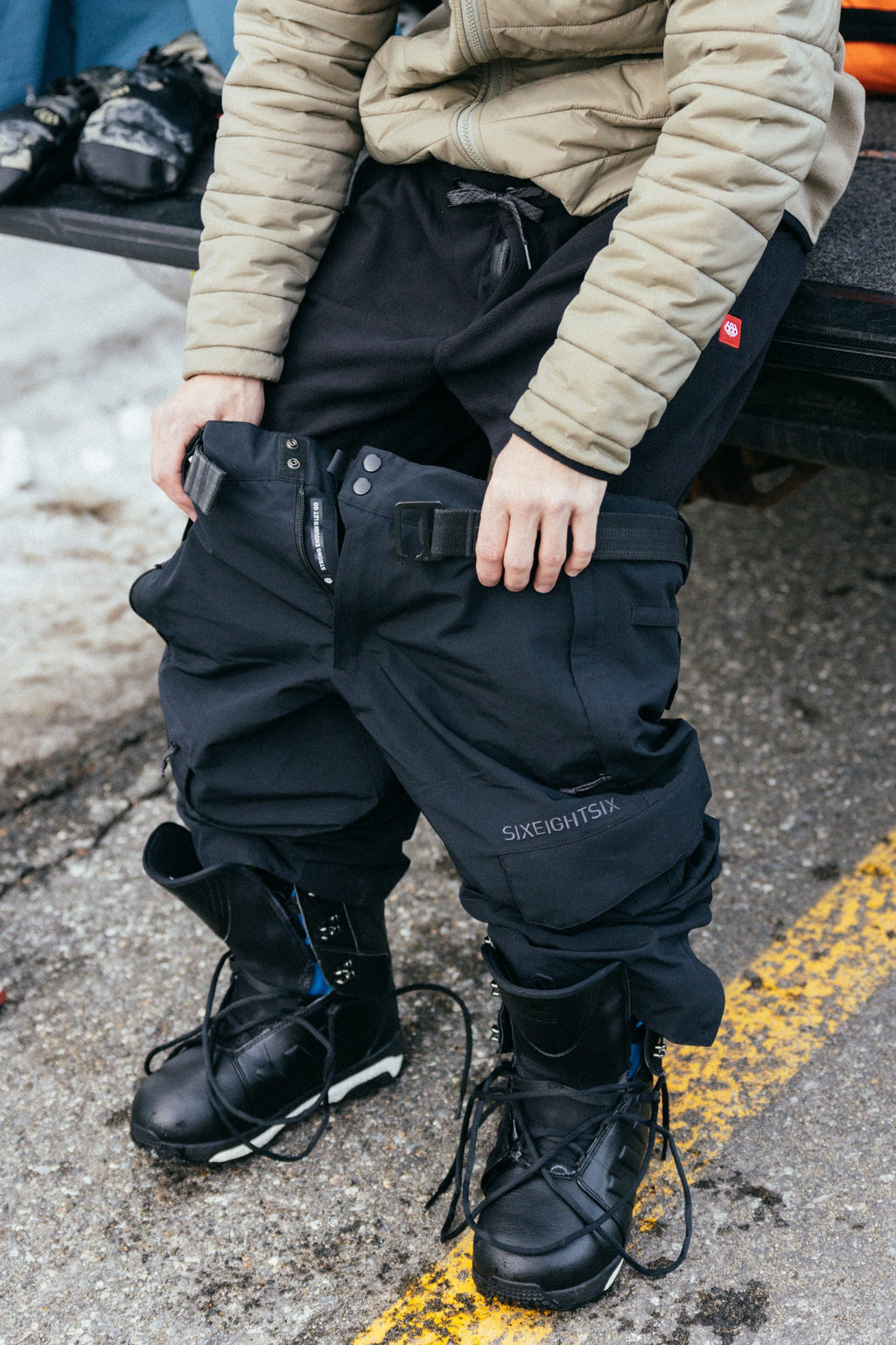 A person in a beige jacket and black snow boots pulls on 686 Men's SMARTY® 3-in-1 Cargo Pants near a snowy street.