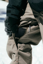 A person in 686 Men's SMARTY® 3-in-1 Cargo Pant stands on snow, unzipping a vent to reveal mesh, highlighting its versatile wear—thanks to the removable sweatpant liner from 686.