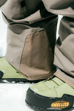 Close-up of a person wearing 686 Men's SMARTY® 3-in-1 Cargo Pant with removable liner and green Deeluxe snowboard boots, standing on snow.