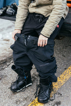 A person sits on a tailgate in a tan jacket and black boots, pulling on 686 Men's SMARTY® 3-in-1 Cargo Pants, which feature a removable liner for versatile wear. Snow and equipment are visible in the background.