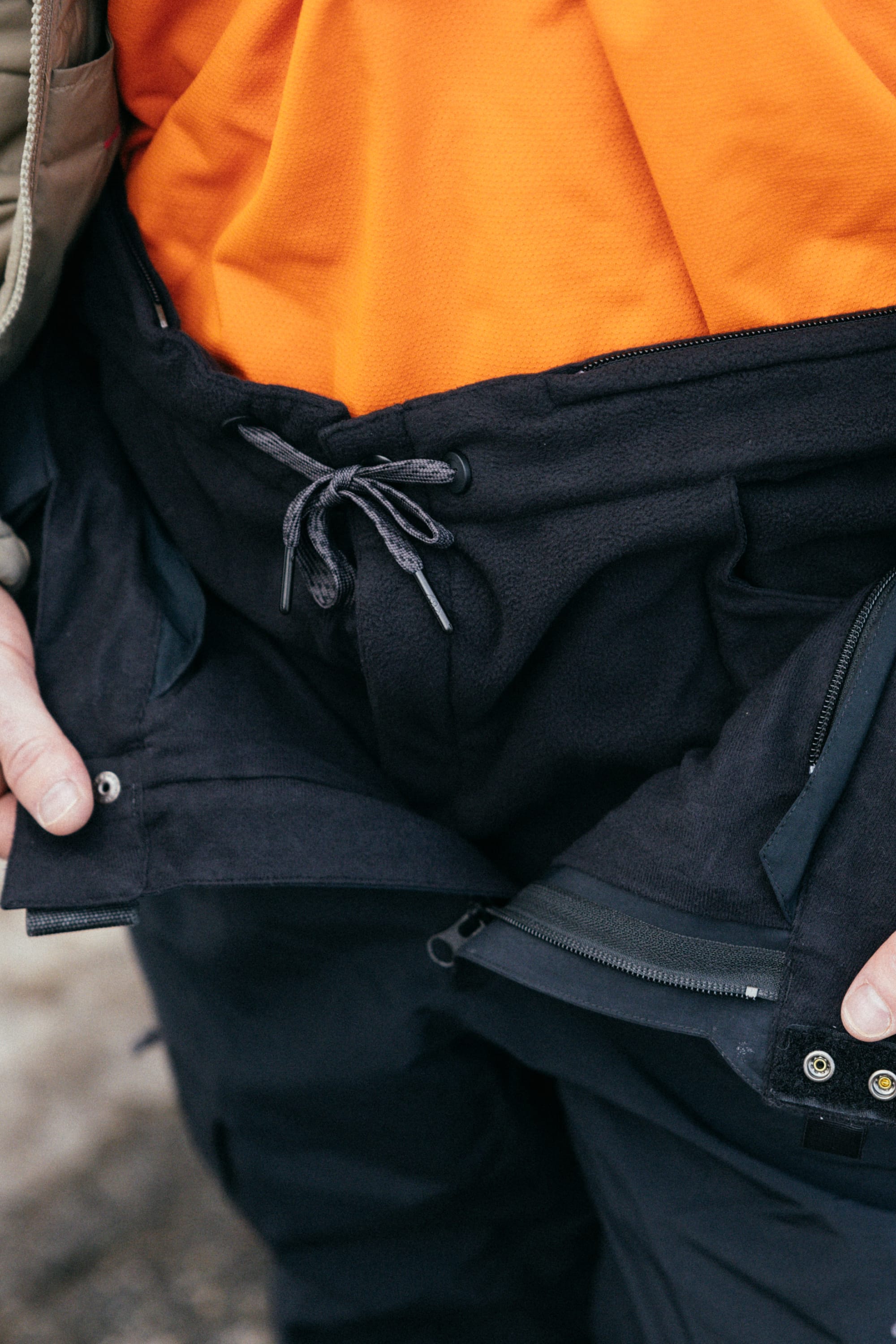 A person wears the 686 Mens SMARTY 3-in-1 Cargo Pant, featuring a drawstring, zipper, and removable sweatpant liner—shown from the waist down with hands near the waistband to display its versatile 3-in-1 wear options.