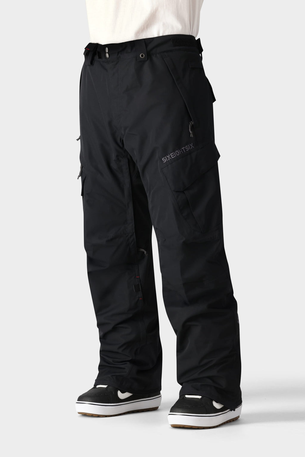 A person wears 686 Men's SMARTY® 3-in-1 Cargo Pant—loose black cargo pants with zippered pockets and a removable sweatpant liner—for three versatile wear options, styled with black and white sneakers and a white shirt. Waist-to-feet view, plain light background.