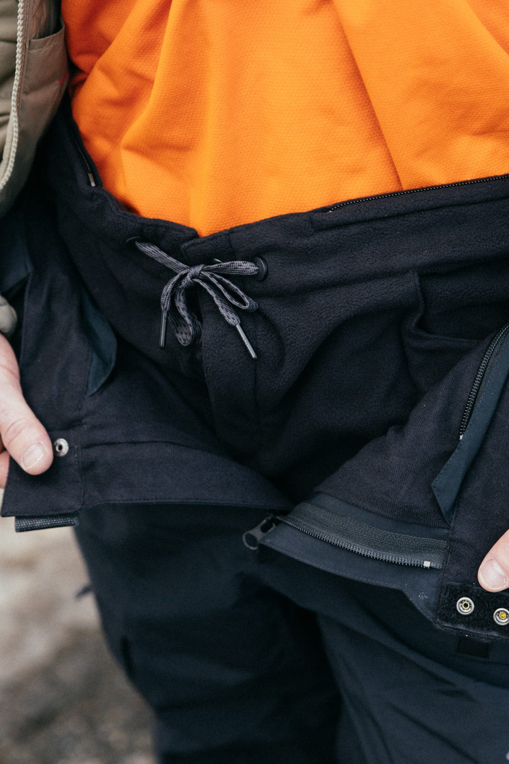 Wearing the 686 Men's SMARTY® 3-in-1 Cargo Pant in black with a drawstring waist, paired with a bright orange shirt and an unzipped black jacket. Their hands adjust the versatile 686 pants outdoors—ideal for three ways to wear.