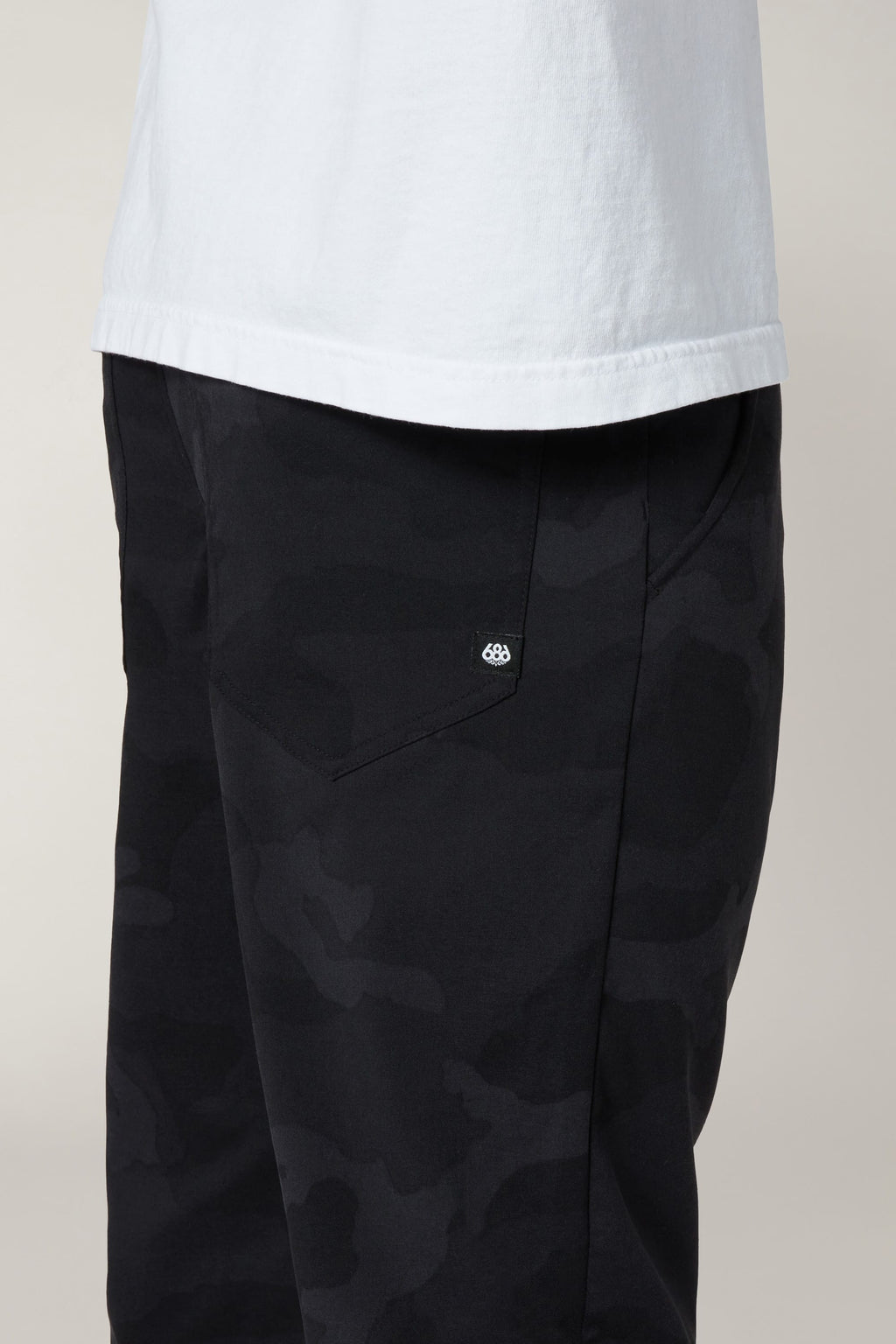 A person shown from the waist down wears a white shirt and 686 Men's Everywhere® Pant - Slim Fit in black camo, highlighting the side pocket with a small white 686 logo.
