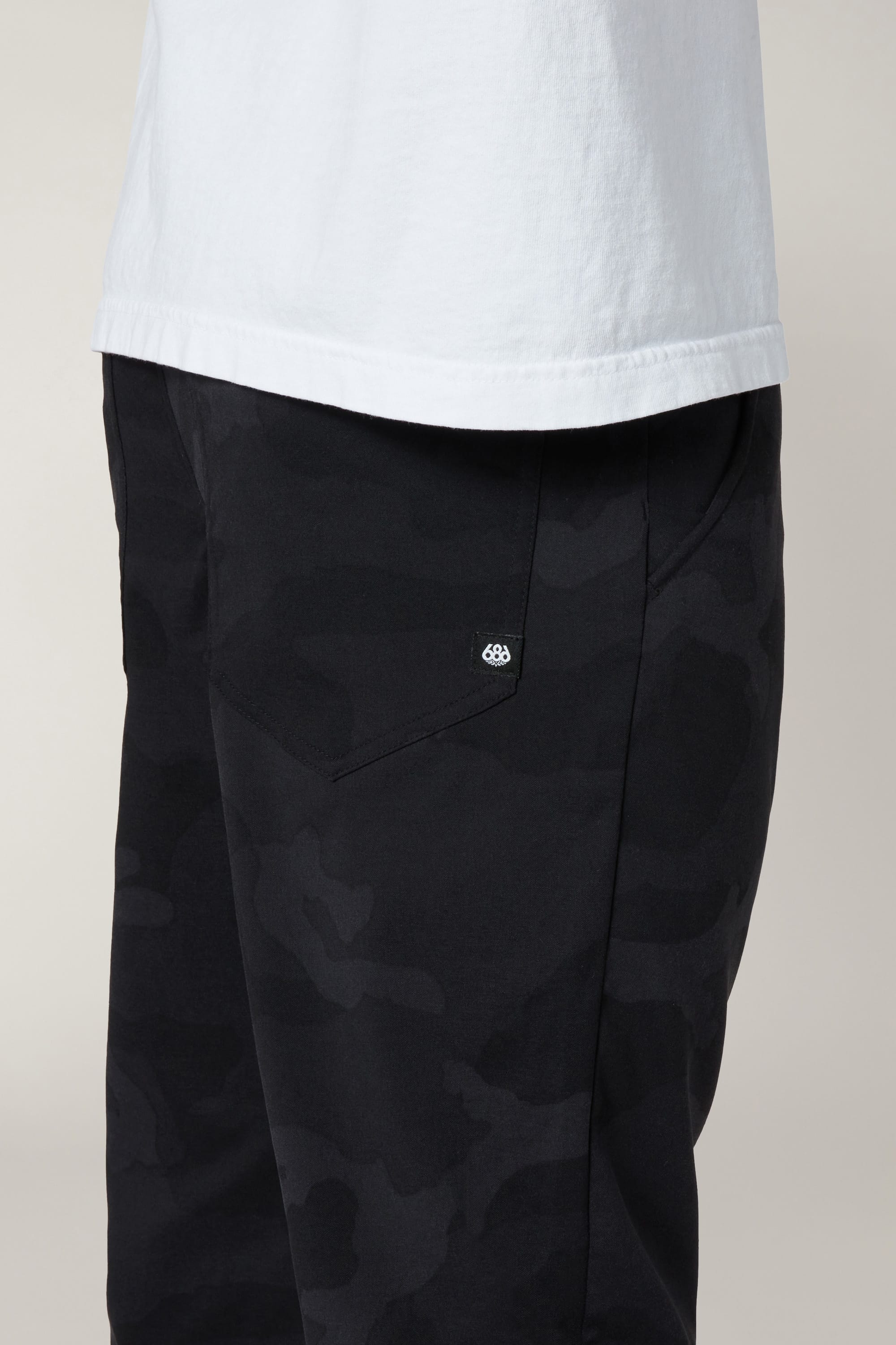 A person shown from the waist down wears a white shirt and 686 Men's Everywhere® Pant - Slim Fit in black camo, highlighting the side pocket with a small white 686 logo.
