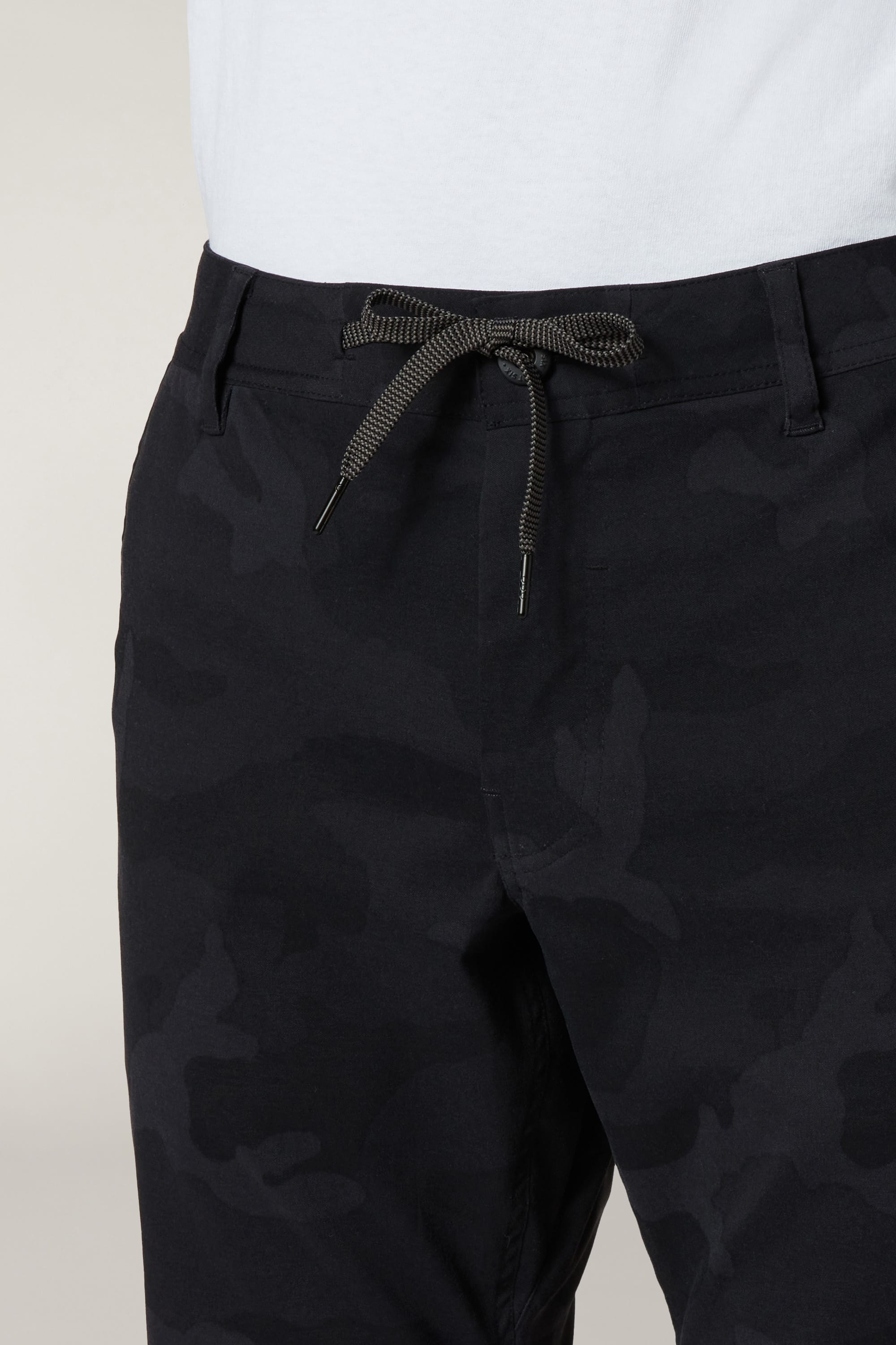 A close-up of a person wearing the 686 Men's Everywhere® Pant - Slim Fit in black camo, featuring a drawstring waist tied in a bow and paired with a white shirt. The pants are breathable and stain-resistant.