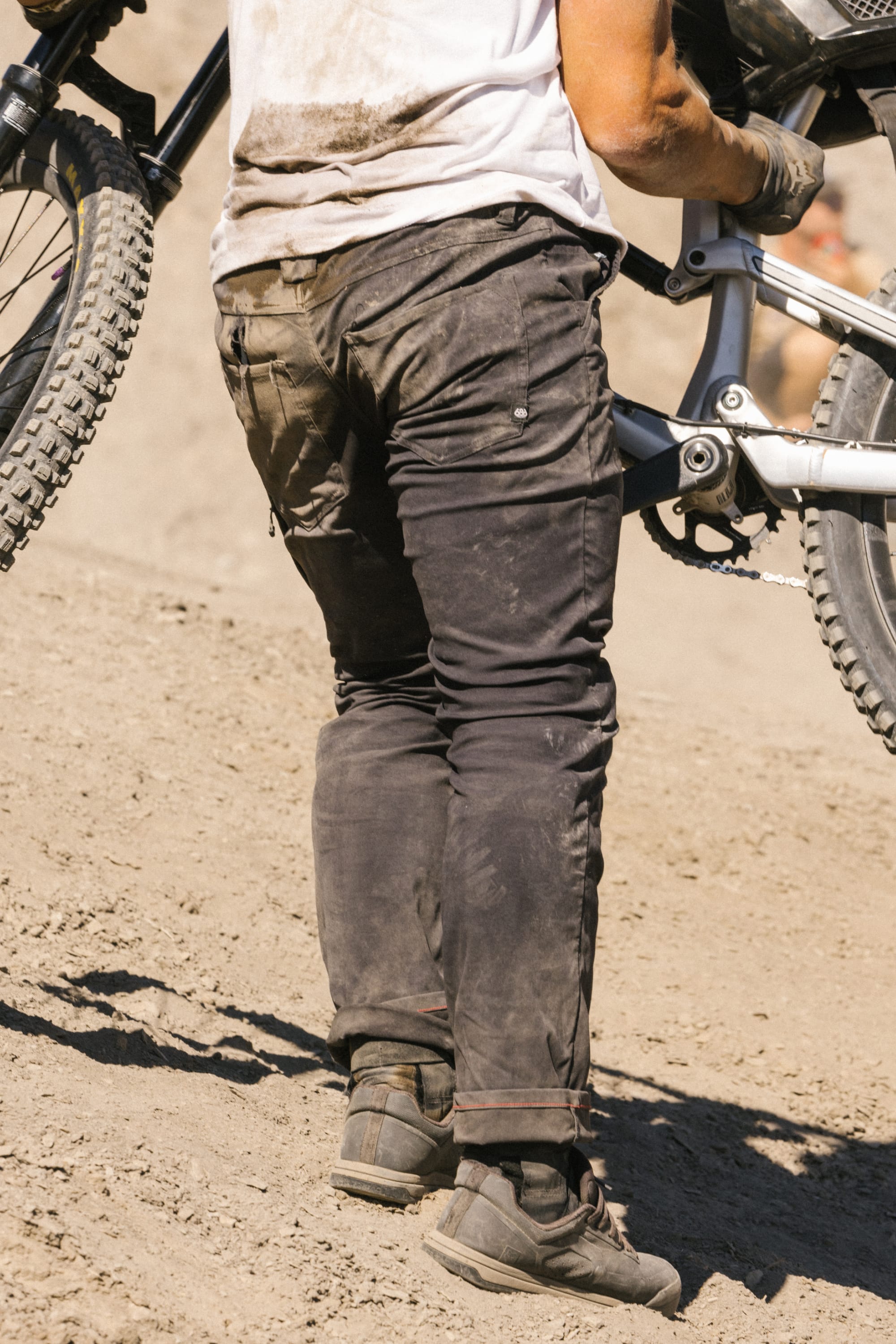 A person wearing 686 Men's Everywhere® Pant - Slim Fit in black and boots stands on a dusty slope, holding a mountain bike by the frame. Only their lower body and part of the bike are visible.