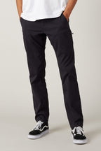 A person wears 686 Men's Everywhere® Pant - Slim Fit in black by 686, paired with a white T-shirt and black-and-white sneakers, standing with hands in pockets against a plain background. The cropped image highlights the pants’ breathable, stylish comfort.
