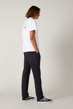 A person with shoulder-length hair stands sideways, hands in pockets, wearing a white graphic-back tee, 686 Men's Everywhere® Pant - Slim Fit, and black Vans sneakers against a plain light background.