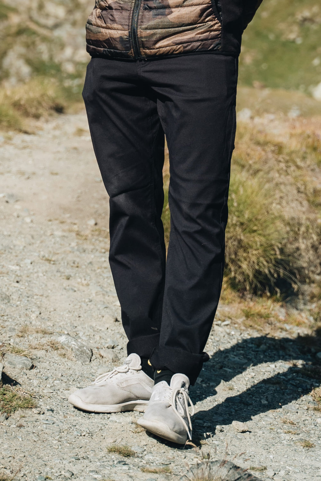 A person stands on a rocky path with green grass nearby, wearing the 686 Men's Everywhere® Pant - Slim Fit in black, along with a brown jacket and light sneakers. The upper body is mostly out of frame.