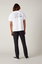 A person with shoulder-length hair stands facing away, wearing a white T-shirt with a black graphic on the back, 686 Men's Everywhere® Pant - Slim Fit by 686, and black shoes, against a plain light background.